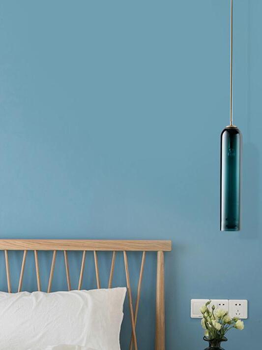 Blue-green glass tube pendant beside a wooden bedhead against a blue wall, with flowers and wall outlets below
