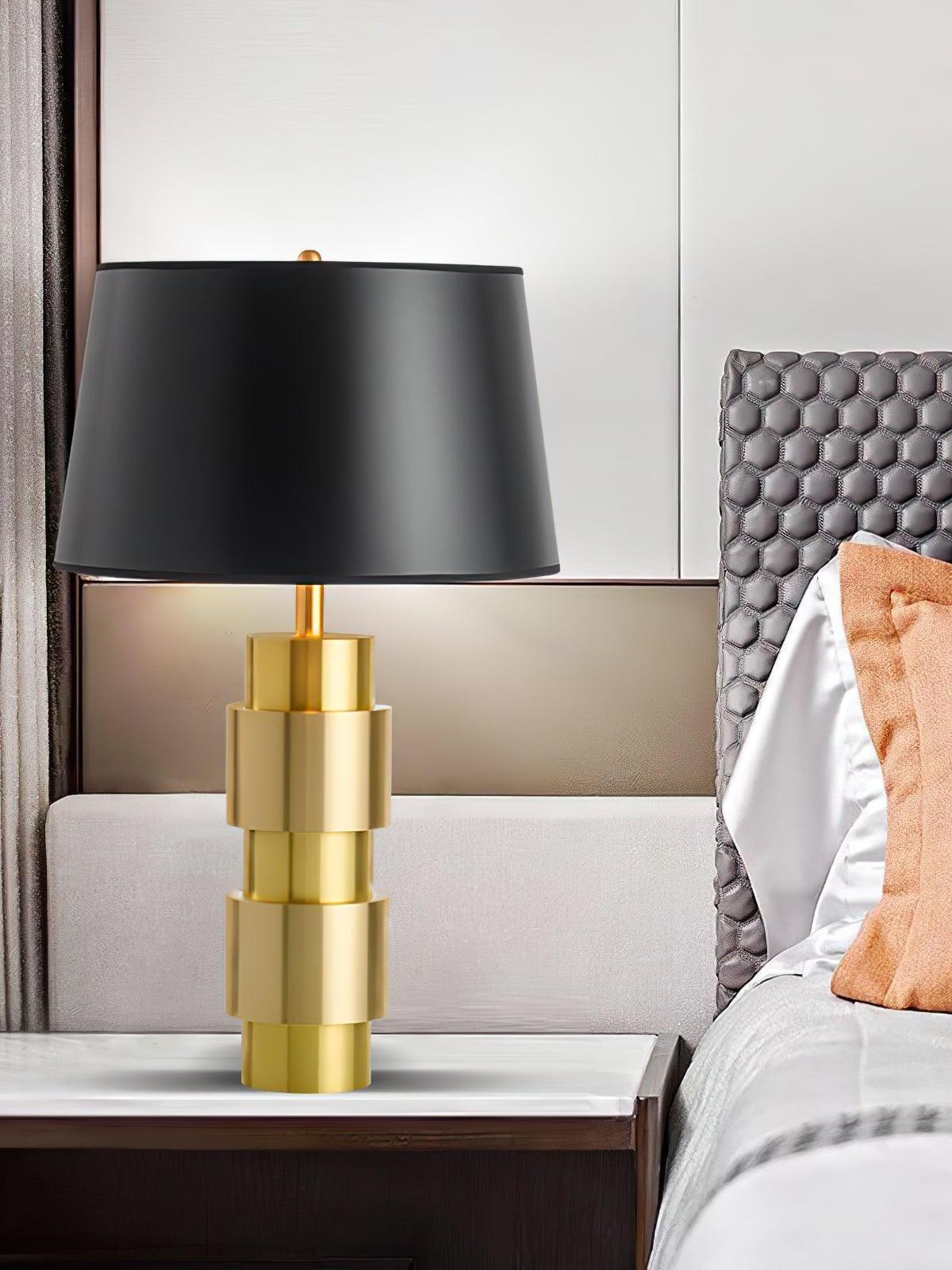 Black shade and stepped gold-tone cylindrical base beside a padded headboard and pale pillow