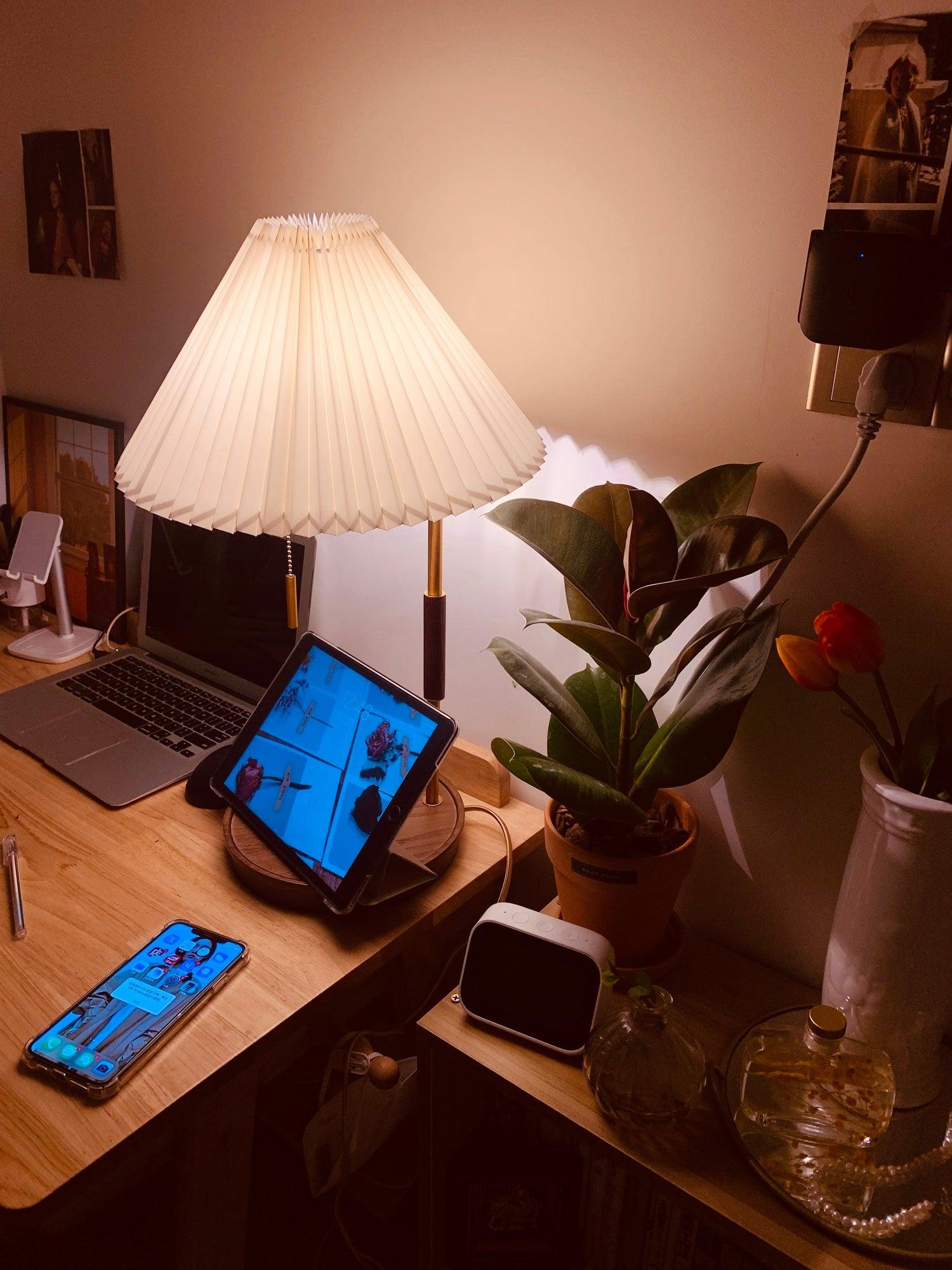 Lit pleated table lamp on a desk with a tablet, phones and a leafy plant nearby