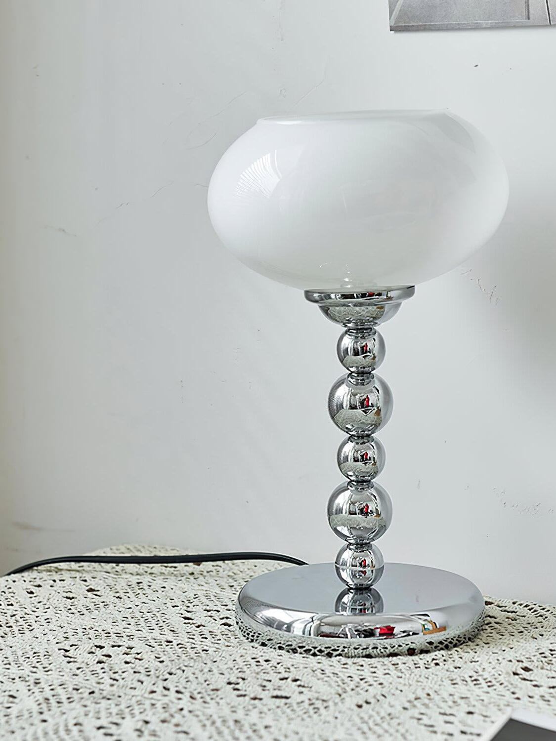 White oval glass table lamp with a rounded chrome stem on a lace-covered surface beside framed art and a black cord