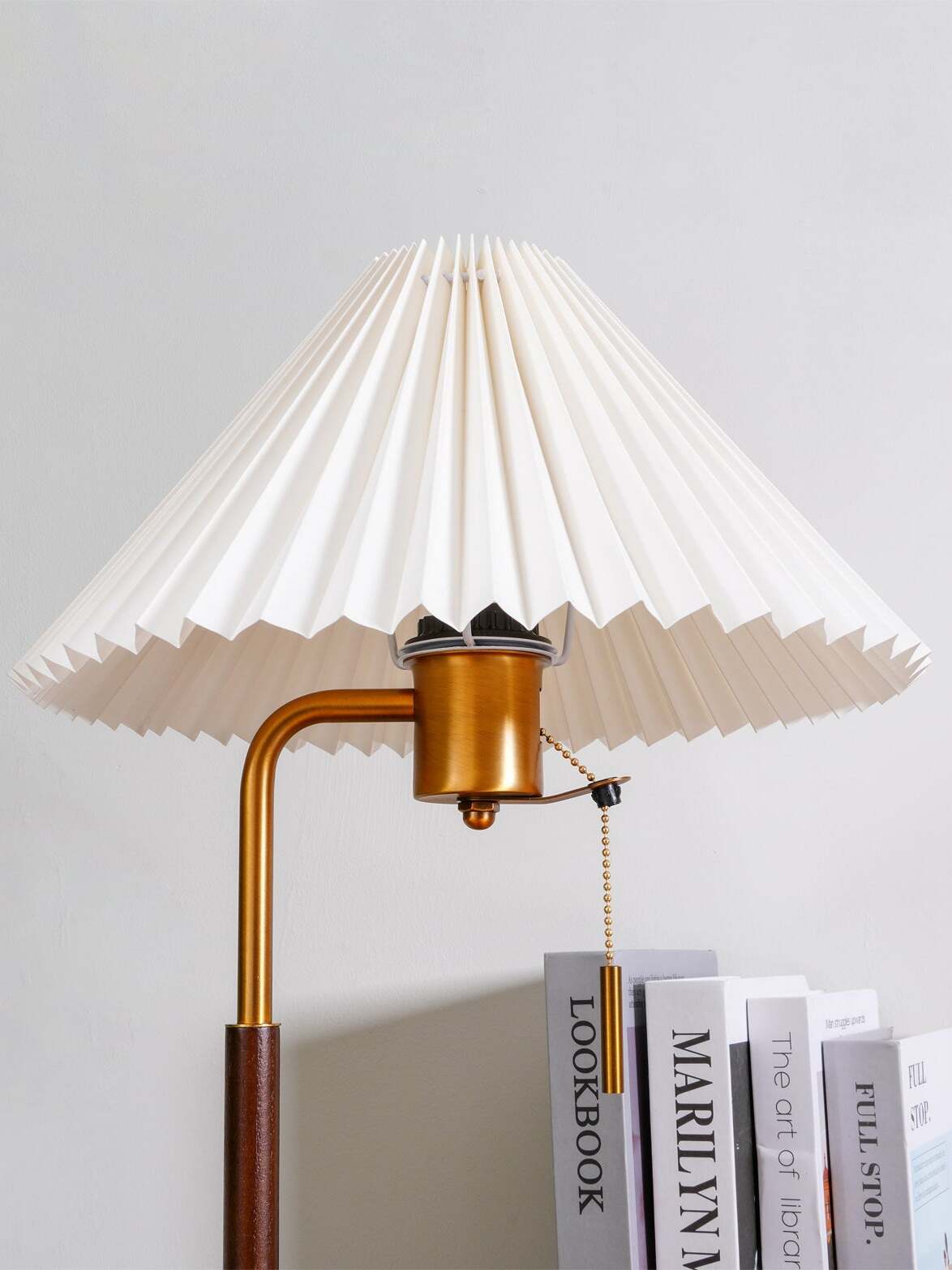 Flared white pleated shade above the curved metal stem and beaded pull chain, with books behind
