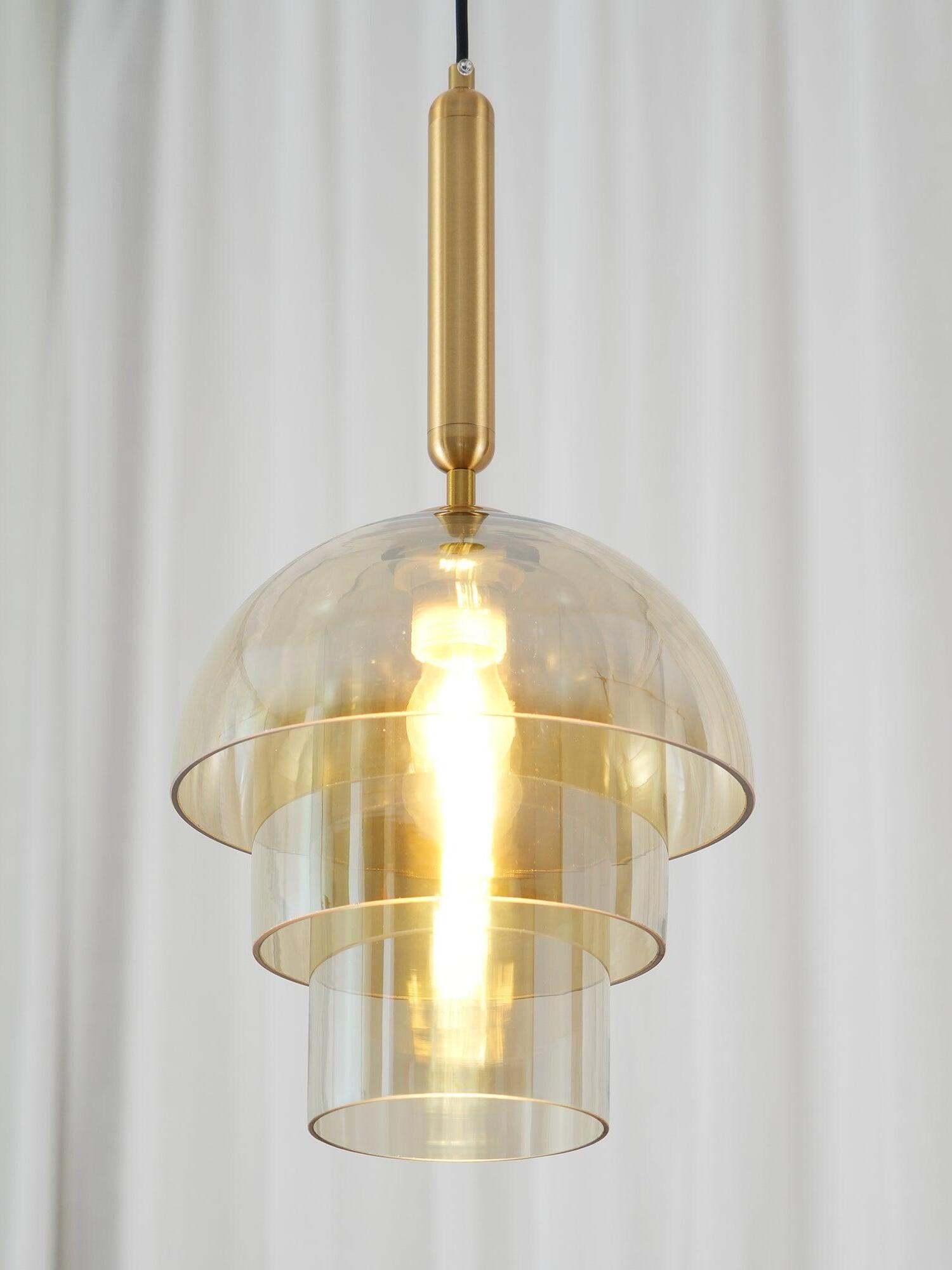 Close view of a lit three-tier glass pendant against white curtains, showing gold-tone top fitting and stepped lower ends