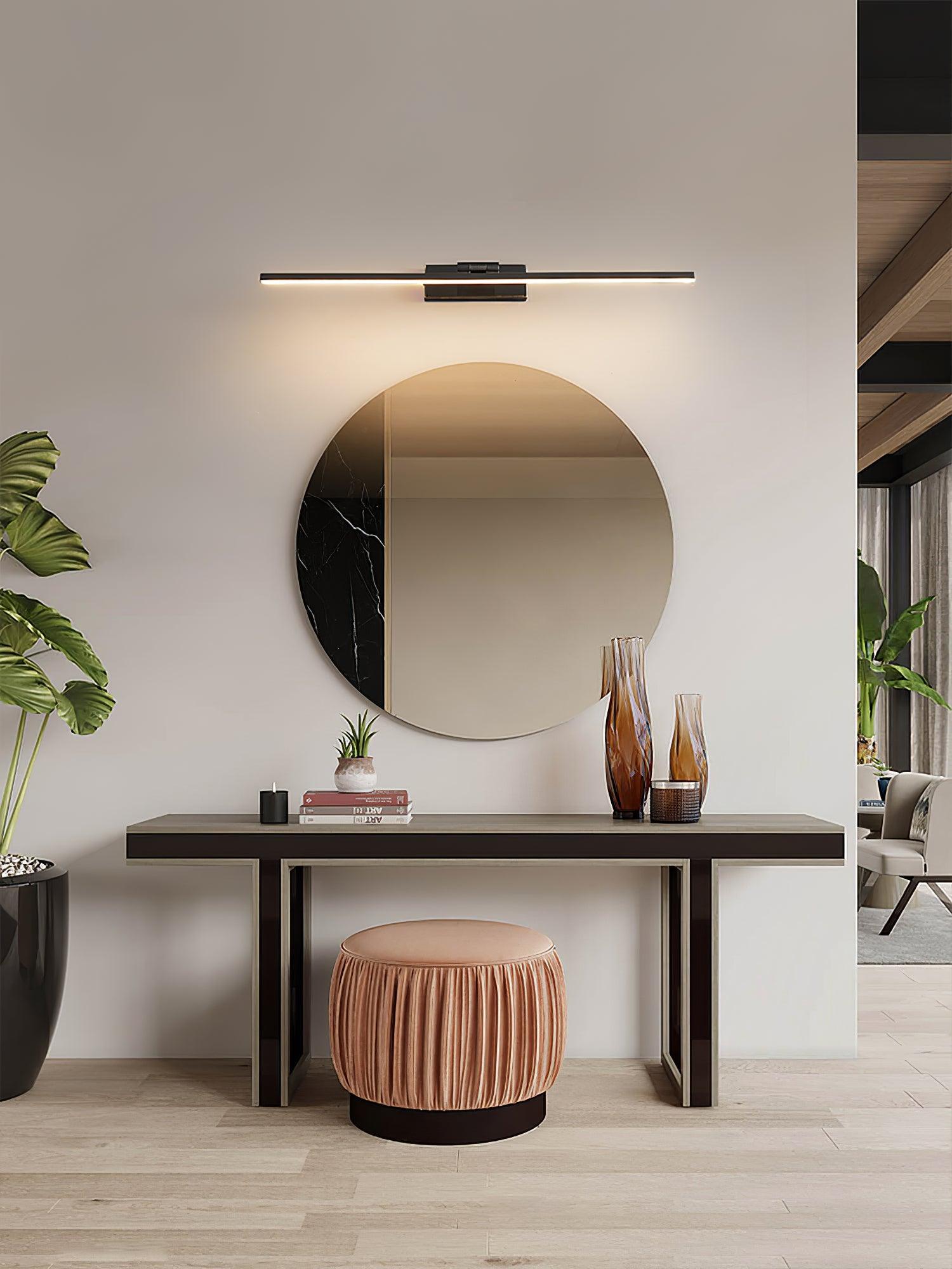 Black linear wall light above round dressing mirror and table with amber vases, books, plants and pink stool