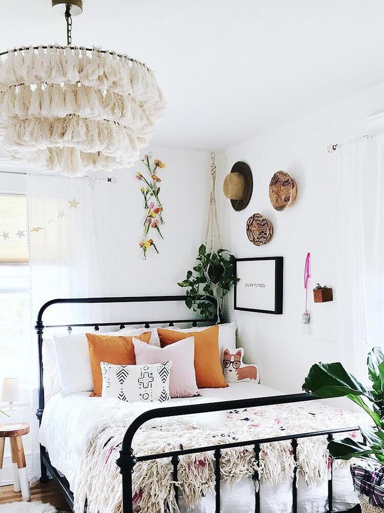 White layered tassel chandelier above a black metal bed with white bedding, orange cushions and hanging plants