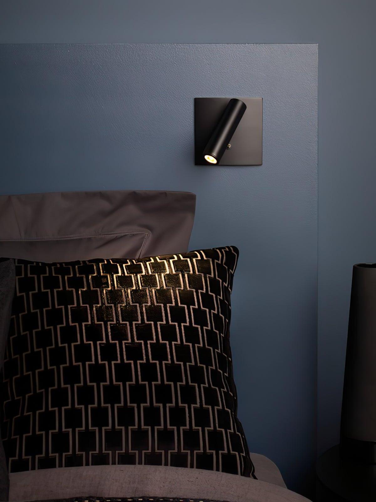 Black Enna cylindrical wall light above bed and black-and-gold patterned pillow against blue wall beside dark table lamp