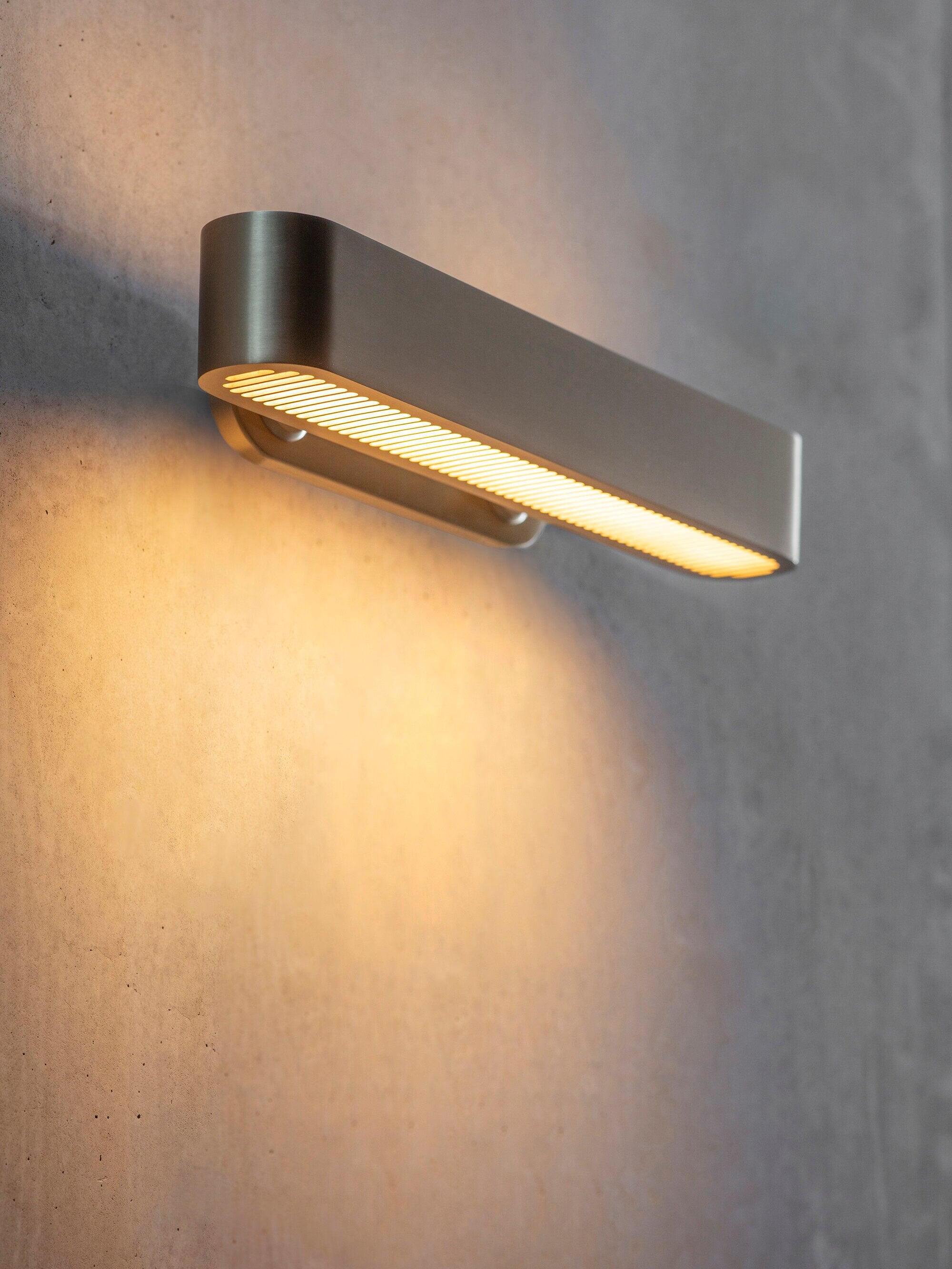 Wider angled view of a horizontal gold-tone wall bar with glowing lower slots and warm wash on a textured wall