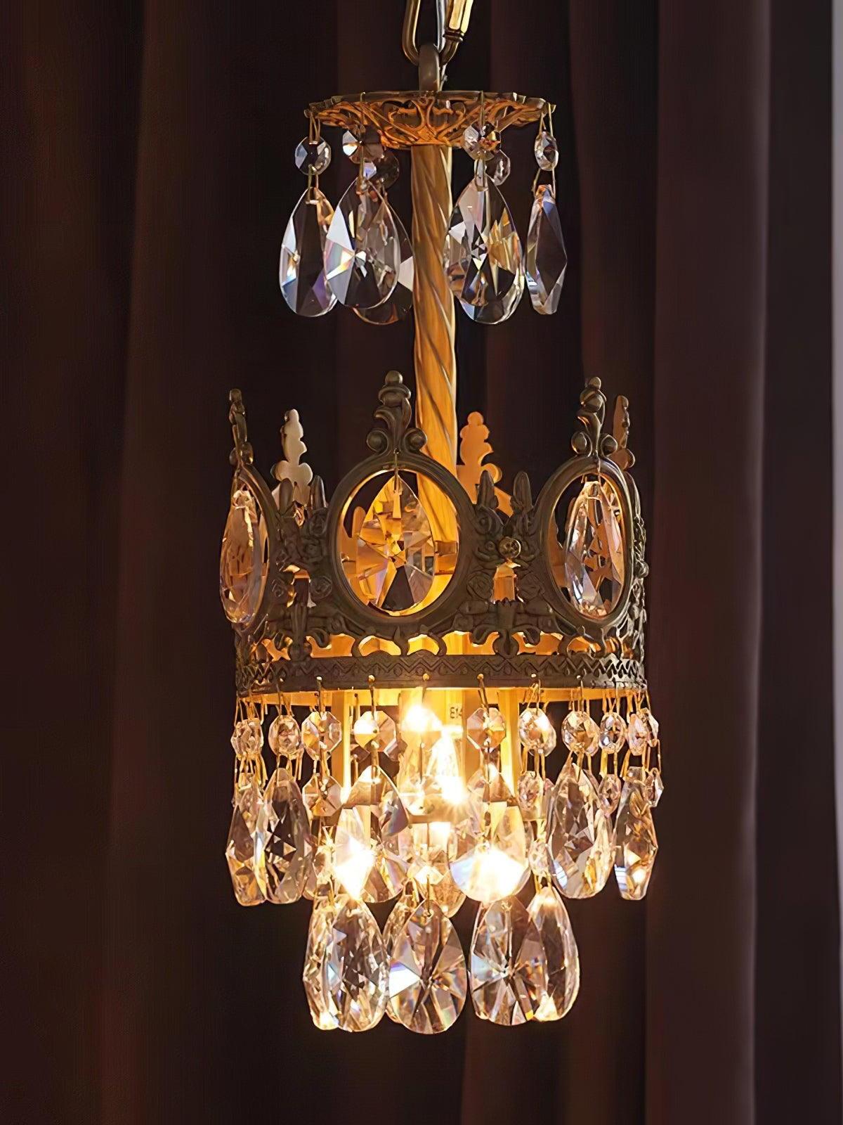 Compact crystal chandelier illuminated against a dark wall, showing its upper disc and lower hanging drops