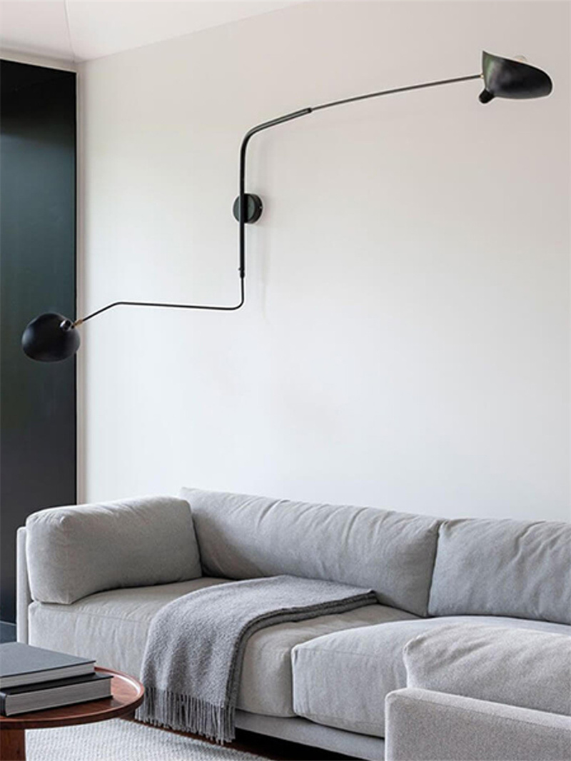 Black two-head wall light with arms extending in opposite directions above a gray sofa and books on a coffee table