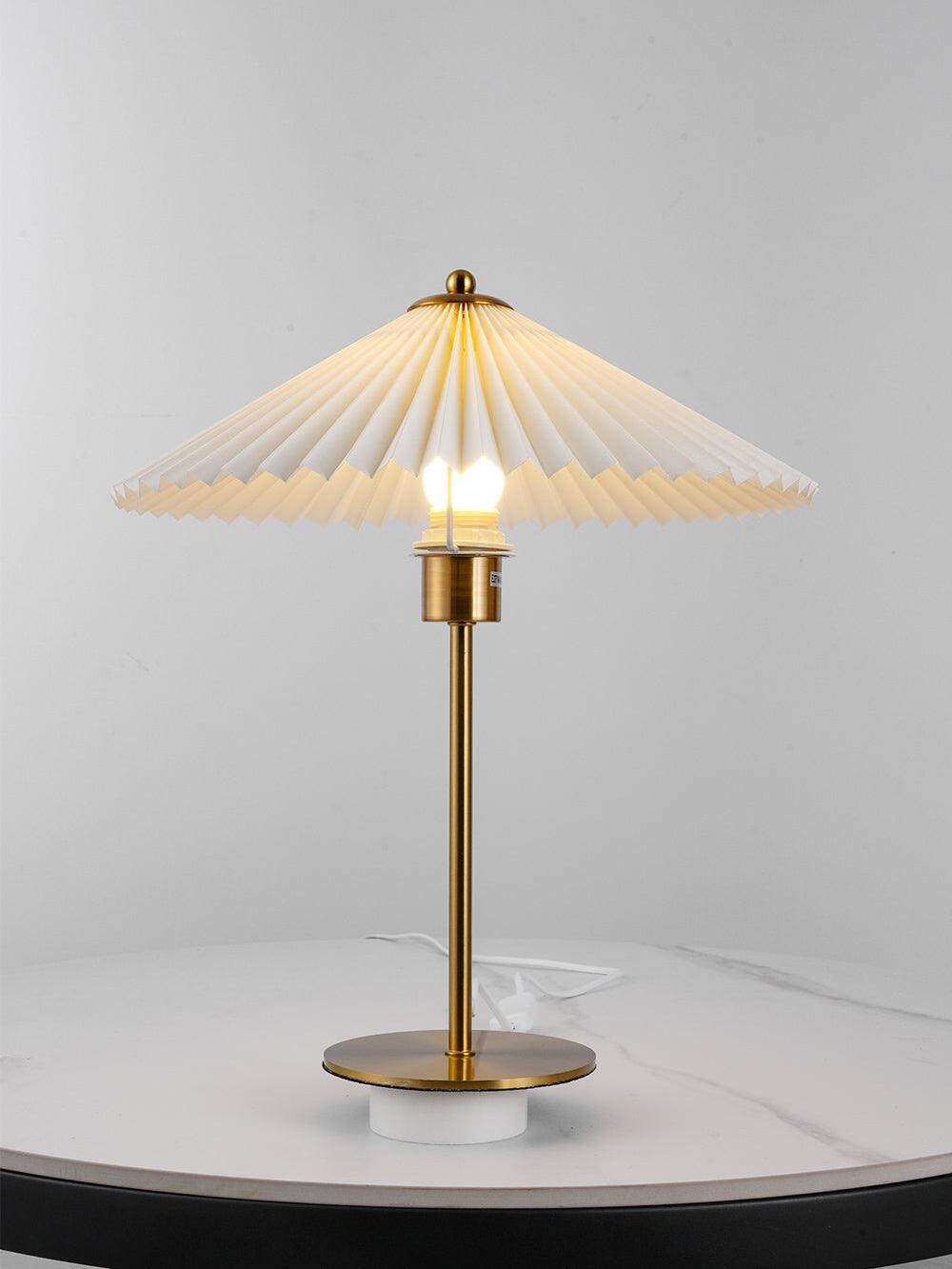 Lit bulb visible beneath the pleated shade above a gold-tone stem and disk base on a white support