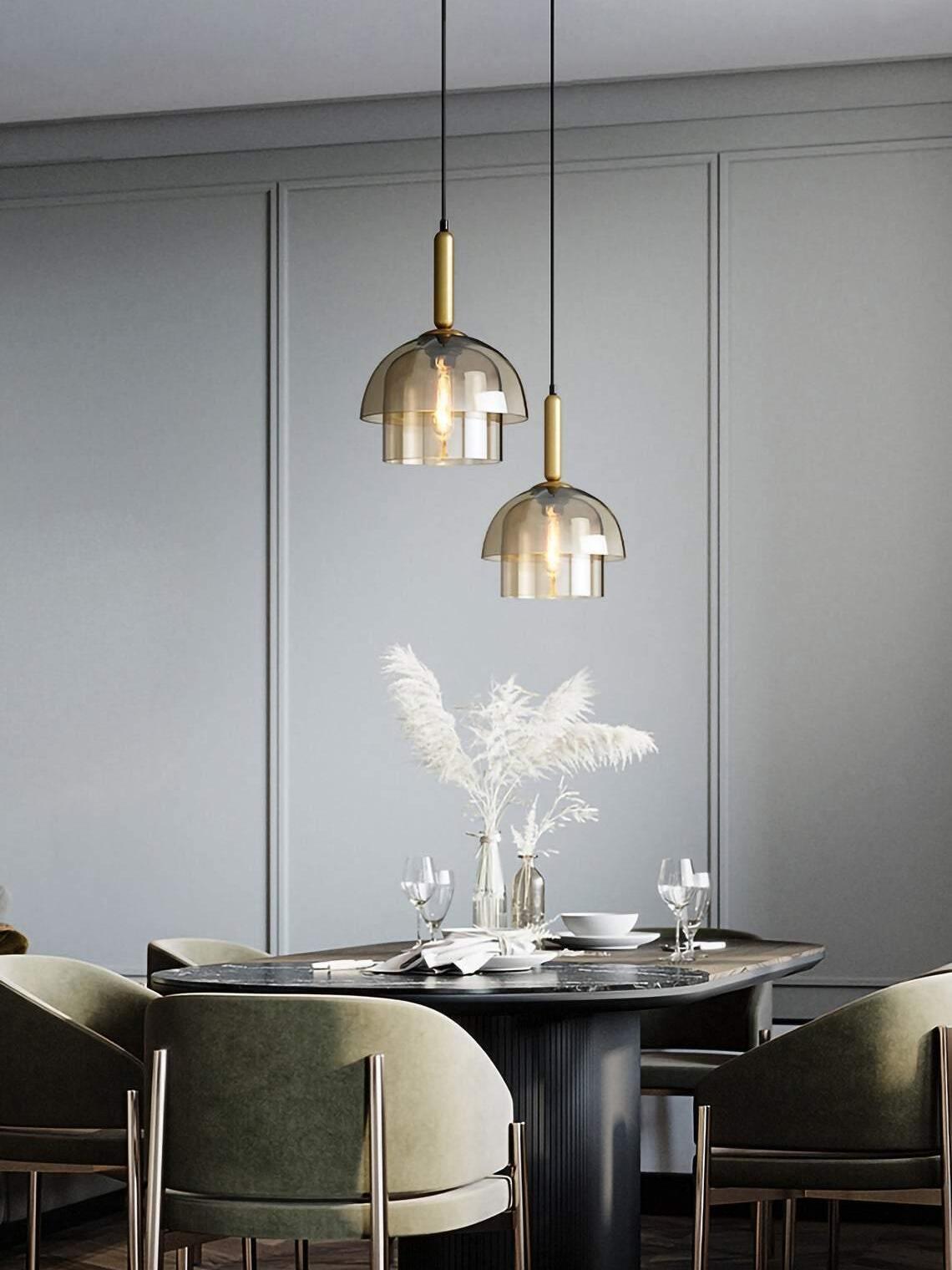 Two two-tier glass pendants over a dining table with white plumes, place settings and green upholstered chairs