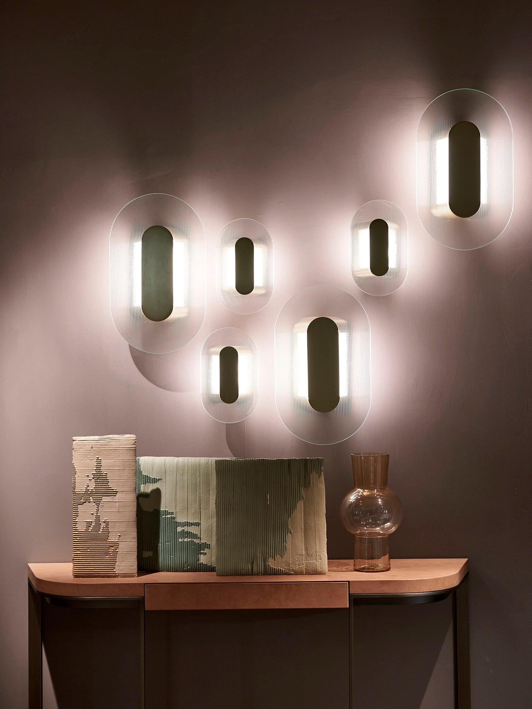 Frontal display of six elongated Button wall lights above a wooden console, decorative boards and glass vase