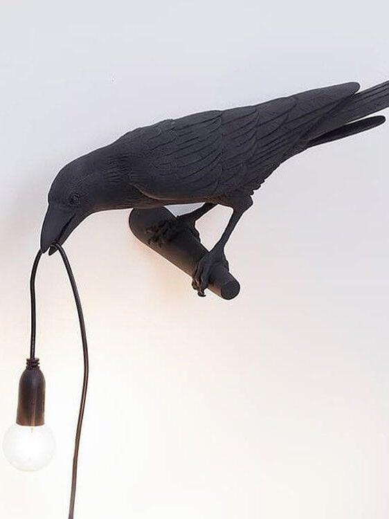 Oblique black bird wall light with a hanging globe bulb, perch and cable against white