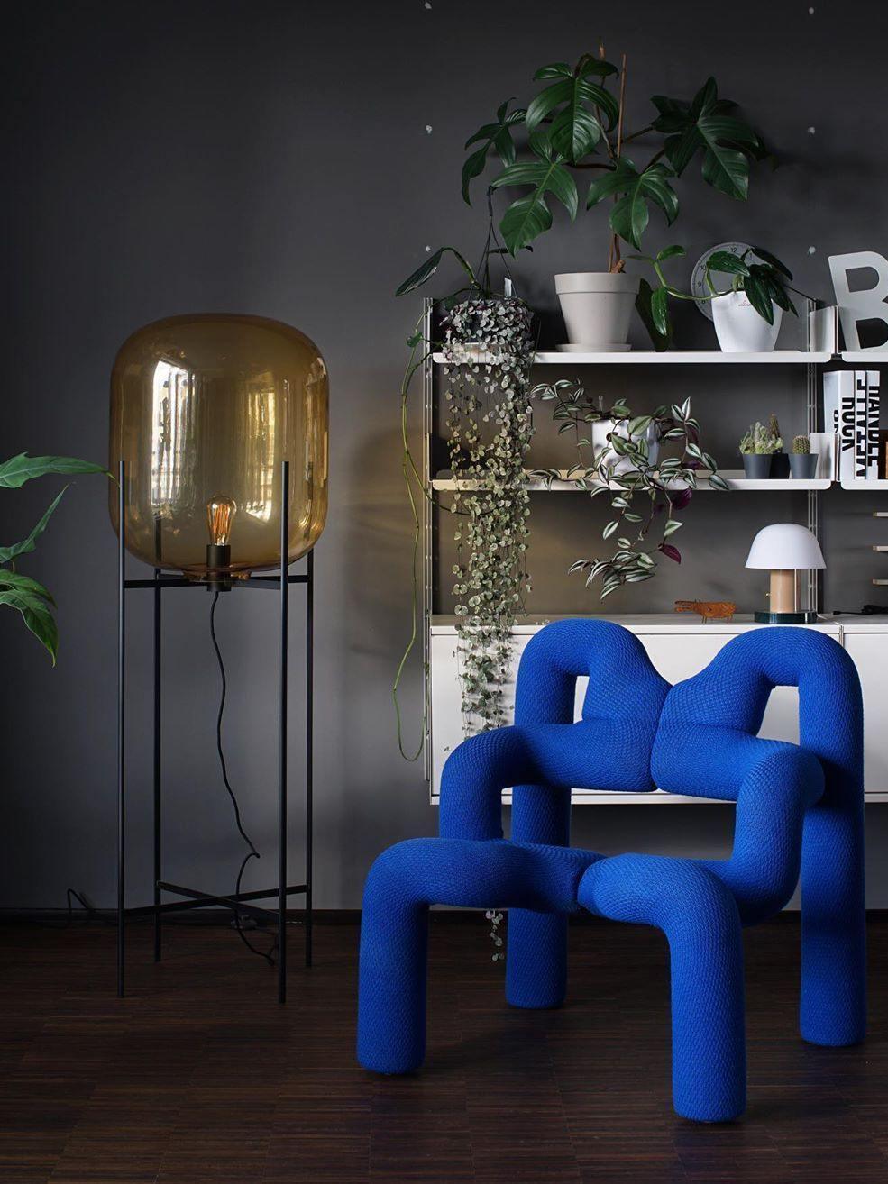Tall amber glass lamp beside a sculptural blue chair and wall shelving.