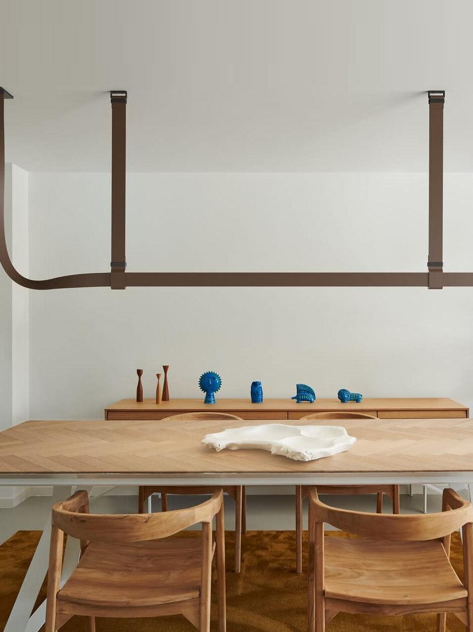 Brown linear pendant above a wood dining table, with a curved strap end at the left and blue figures behind