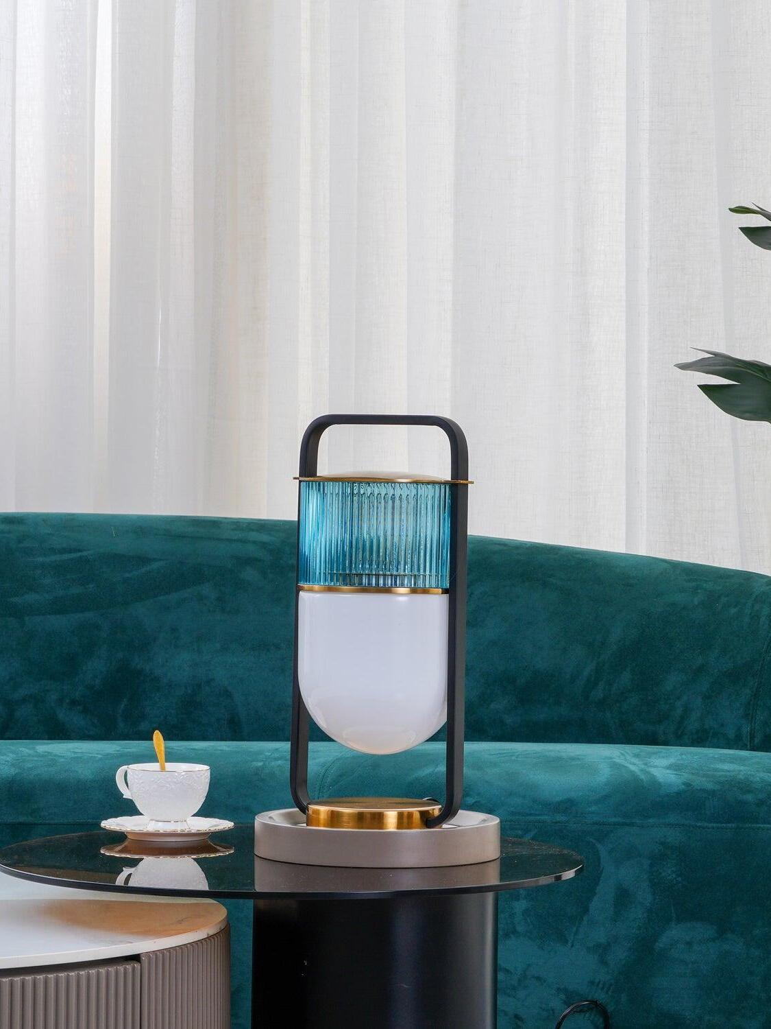Xi lamp on a round glass side table with a cup, white curtains and teal sofa in the background