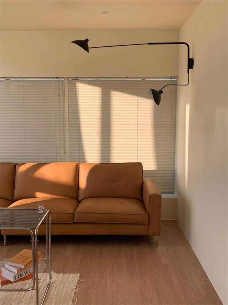 Black two-arm wall light above an ochre sofa beside window blinds and a glass cart, with shades at different heights