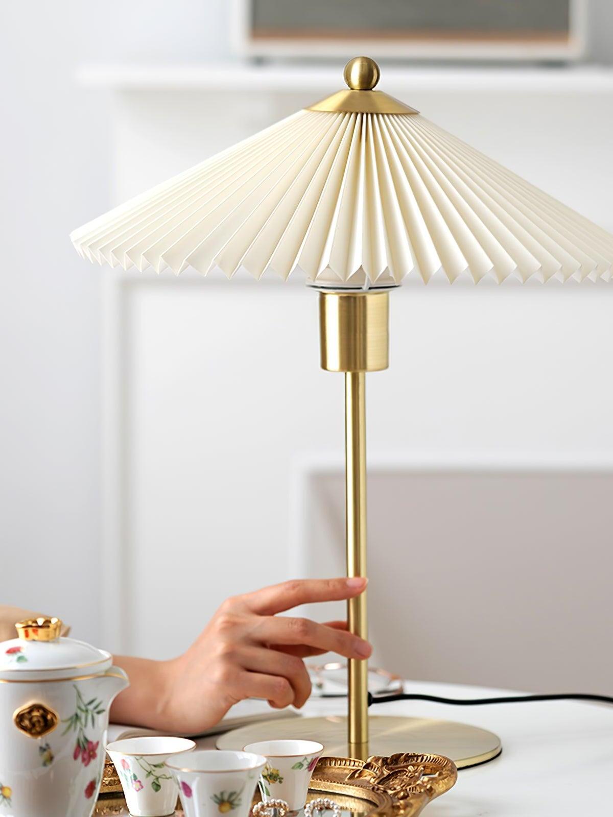 A hand beside the thin gold-tone lamp stem, with a pale pleated shade above and a tea set in front