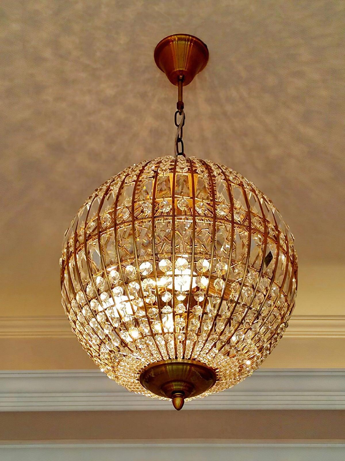 Low view of a lit gold-tone globe cage showing internal lights, clear crystals and a short ceiling chain