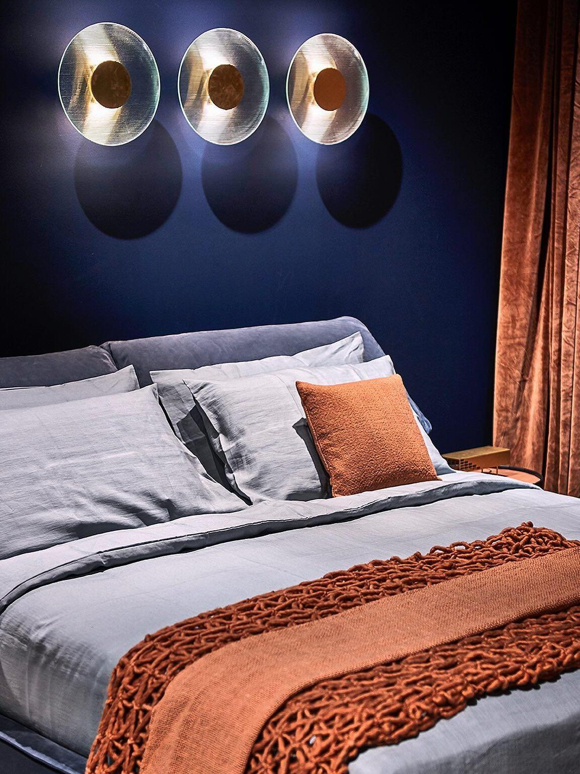 Three round Button wall lights on a blue bedroom wall above pale bedding and an orange throw