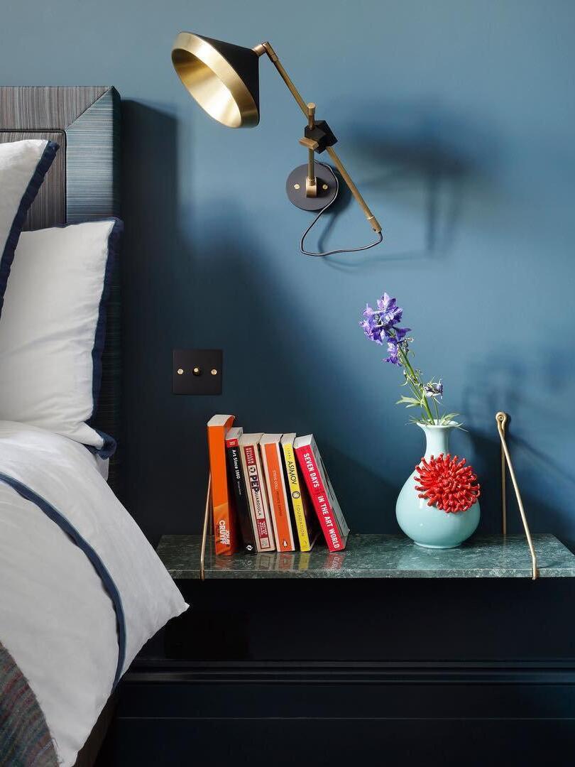 Black cone wall light tilted upward beside a blue bedroom wall, books and a flower vase