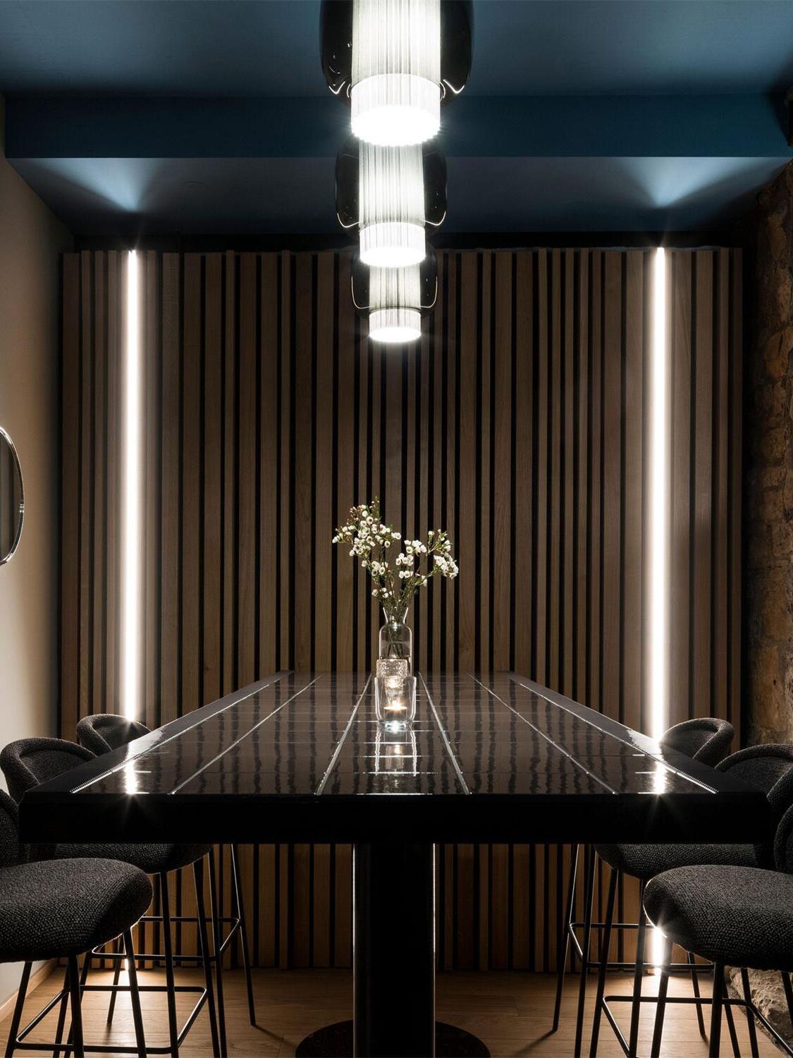Three black and smoke-grey Manila T pendants over a long high table with upholstered stools and a wood-slat wall