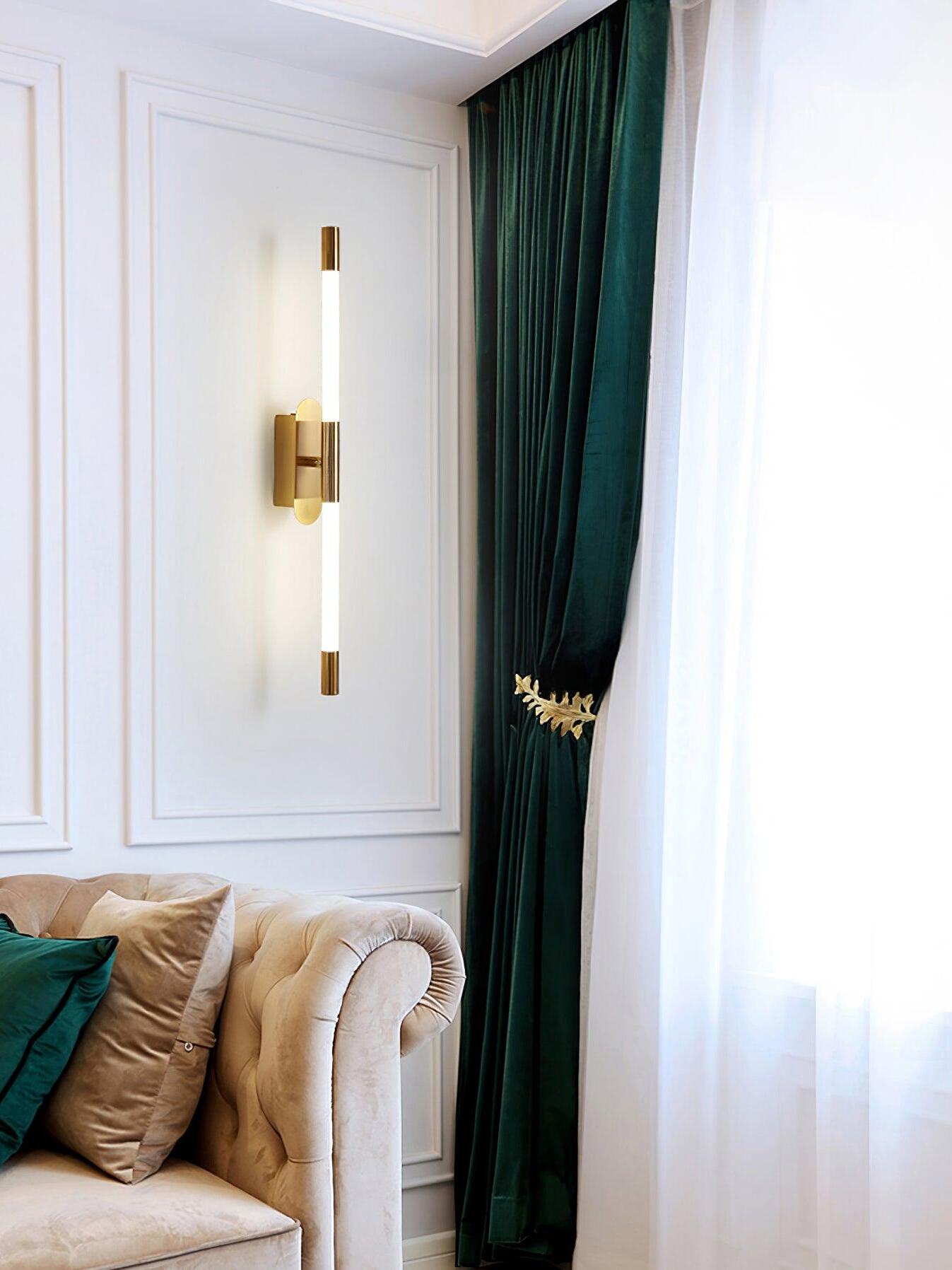 Vertical gold Agnes wall light between a panelled wall and a tall green curtain