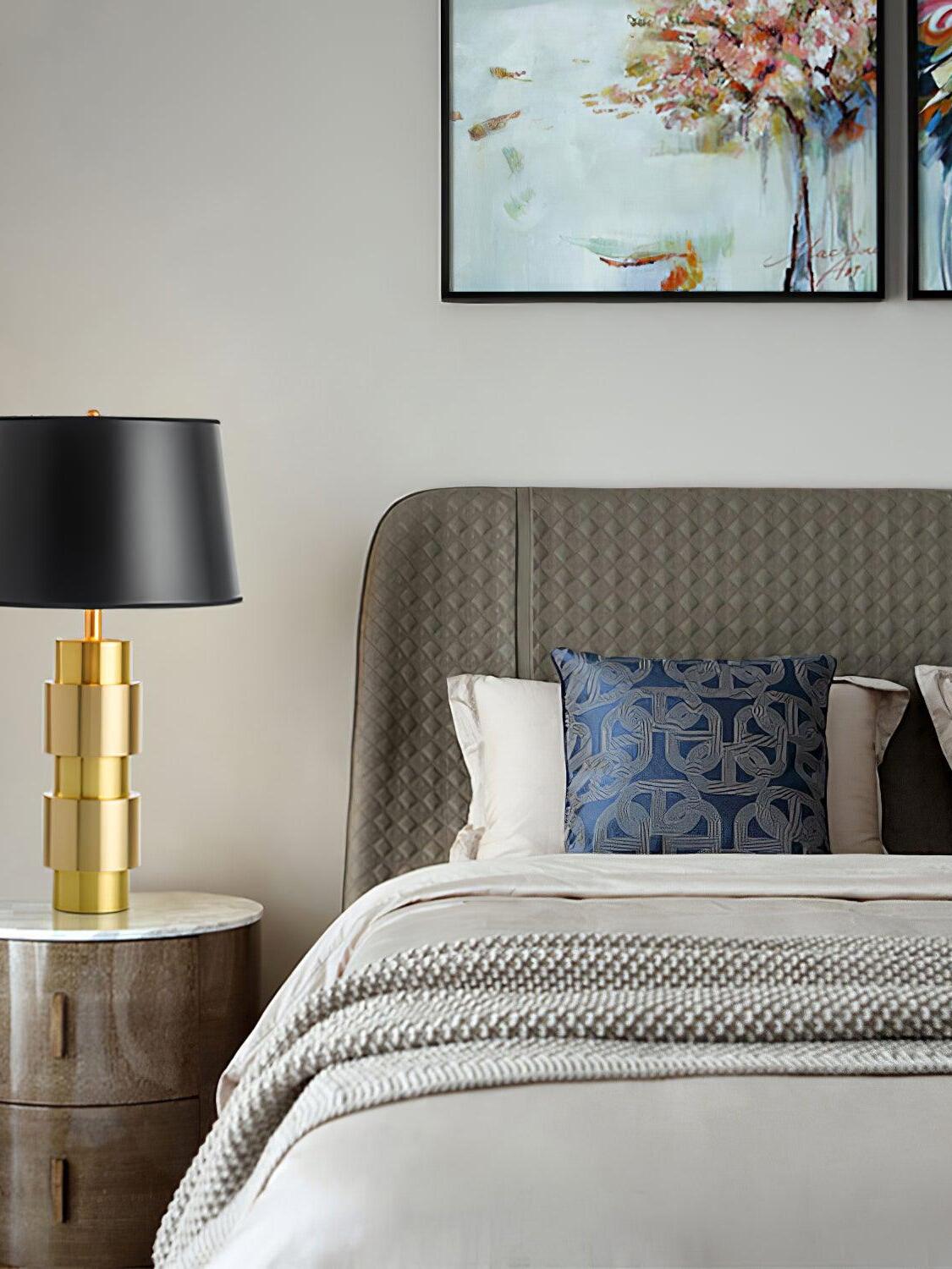 Black-shade lamp on a round bedside table beside patterned bedding and floral artwork