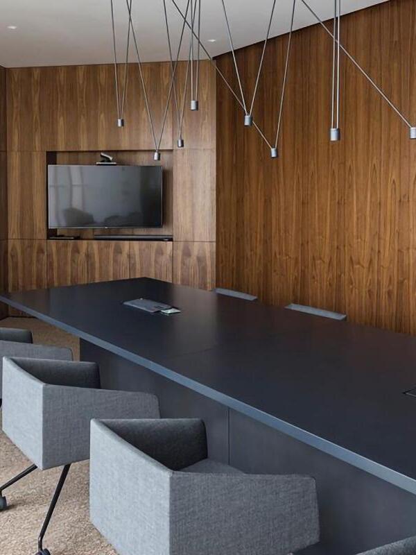 Gray cylindrical lights and crossing rods above a dark conference table and gray chairs before wood paneling