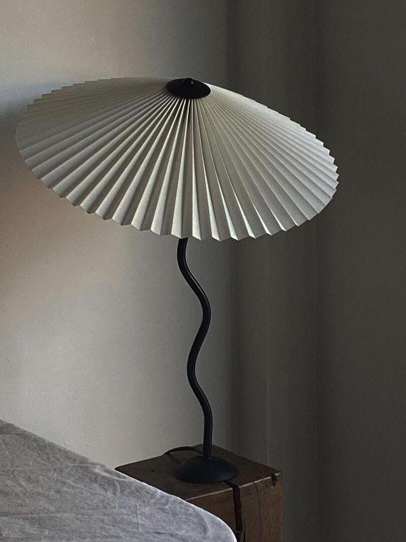 Pale pleated-shade lamp with a black wavy stem on a wooden bedside cube beside gray bedding