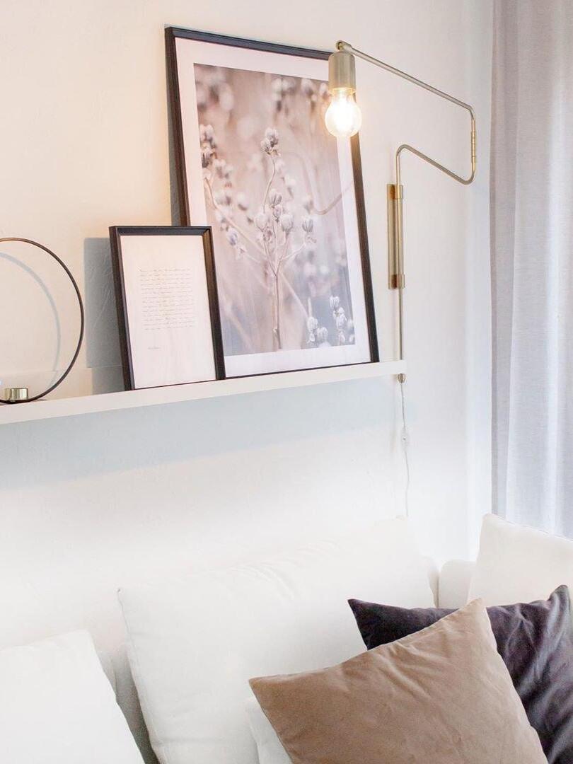 Brass-tone bent-arm wall lamp above a white sofa and picture shelf with framed floral artwork