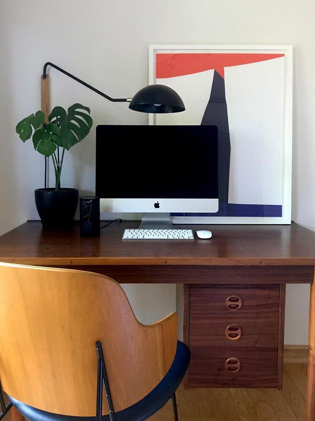 Black dome wall lamp on a light wooden mount above a computer, potted plant and leaning graphic print on a wood desk