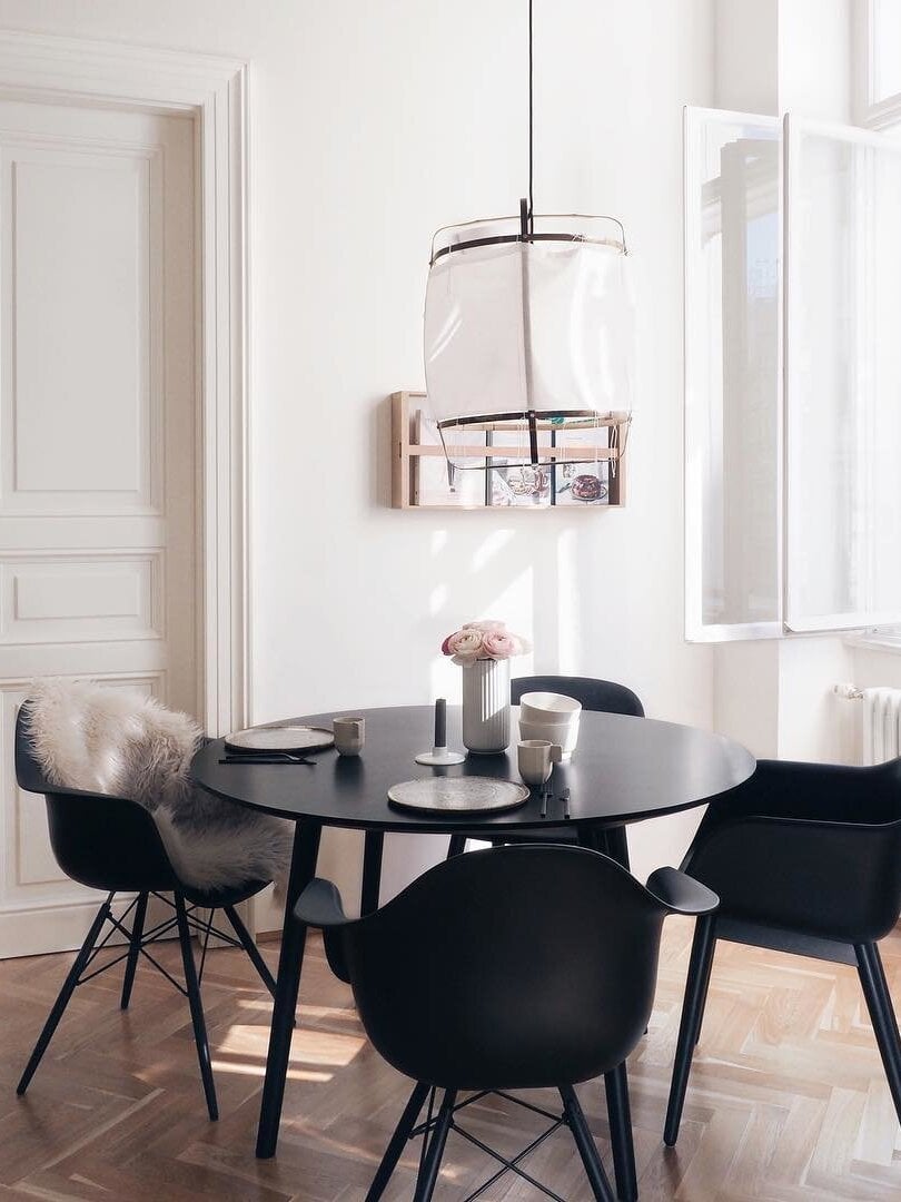 White bamboo lantern pendant over round black dining table and black chairs, beside a white wall and open window