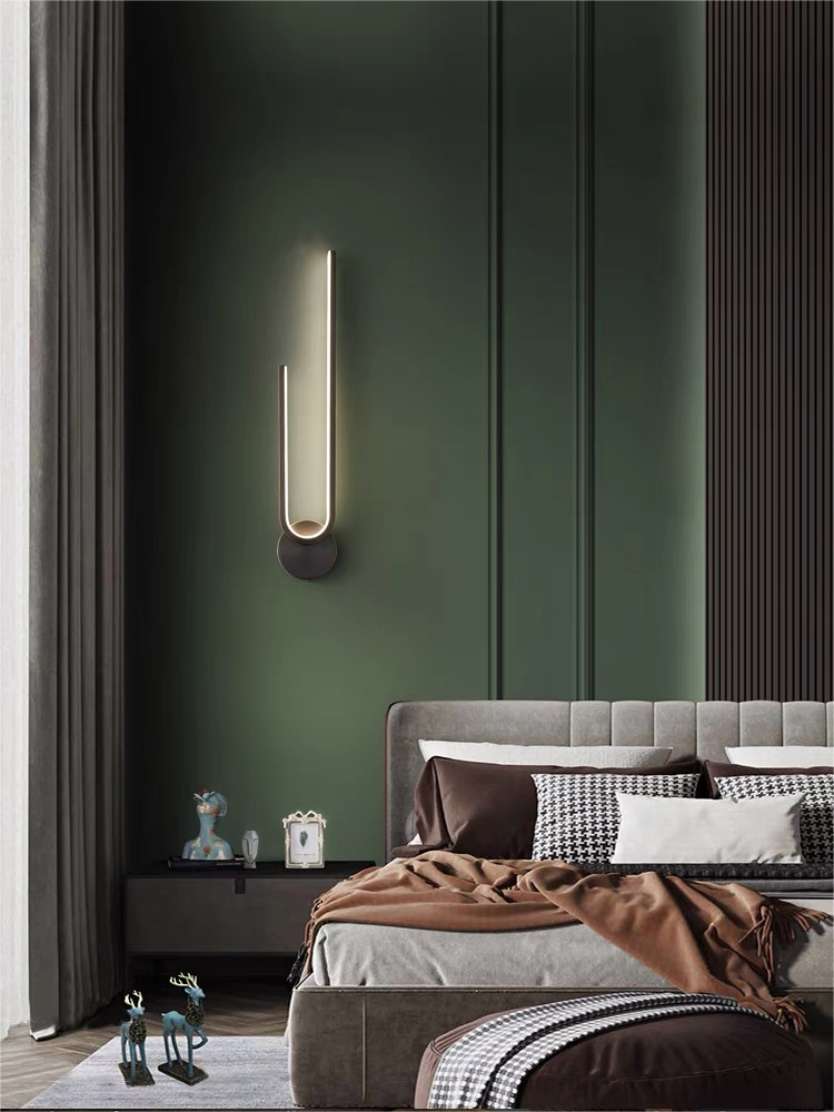 Inverted dark arch wall light with its round mount below the upward arms beside a bed on a green wall