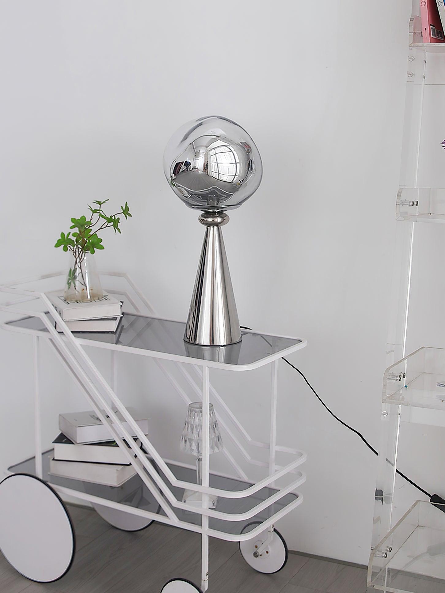Unlit mirrored globe table lamp with a chrome cone base on a white cart beside books and a plant