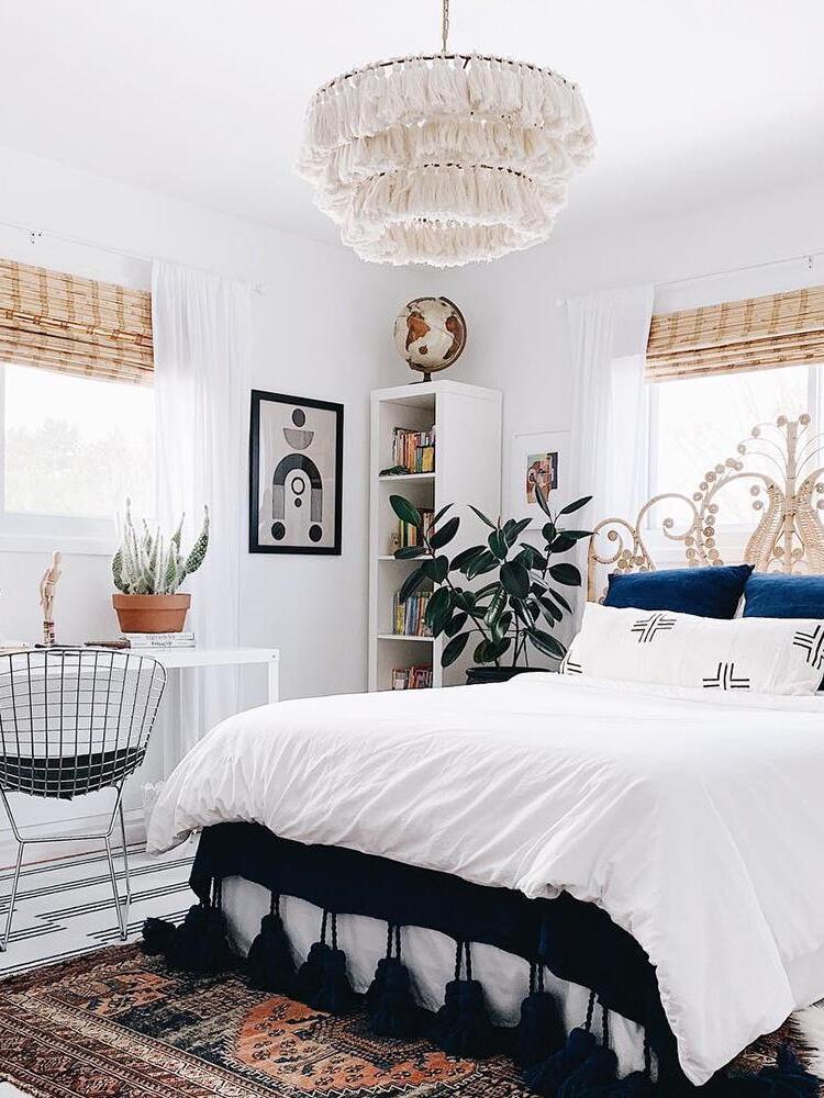 White tassel chandelier in a bright bedroom with blue cushions, a decorative cane headboard, bookcase and woven window blinds
