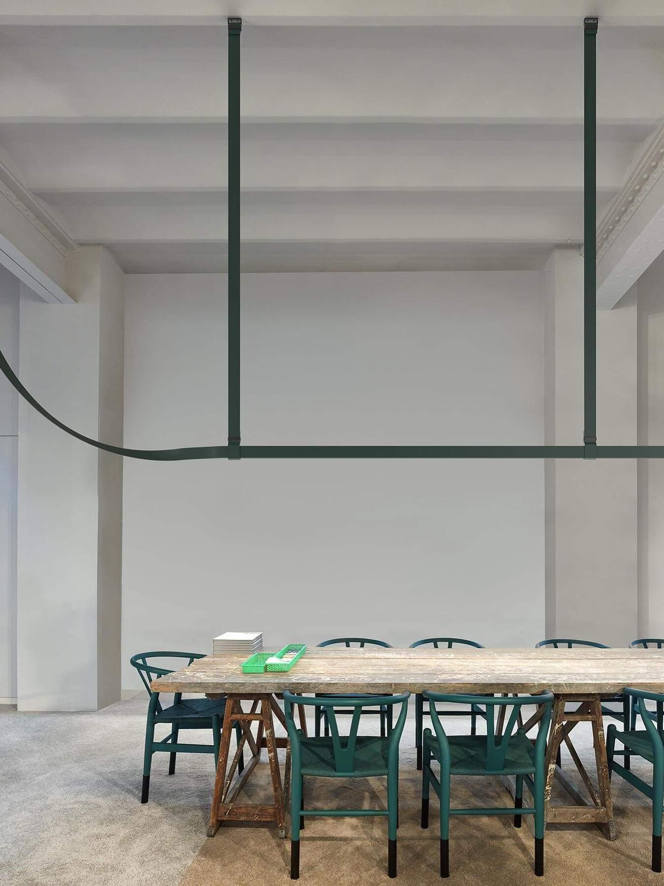 Green long horizontal pendant with two upright straps and a curved left end above a rustic wood table and green chairs