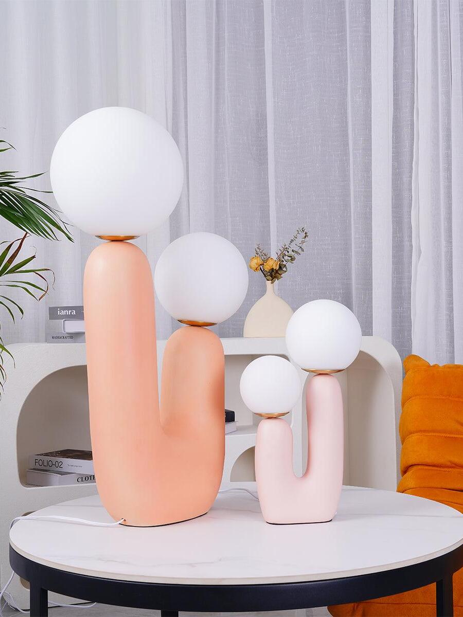 Large peach and smaller pink Oo Rough lamps on round pale table before sculptural shelves, books and orange chair