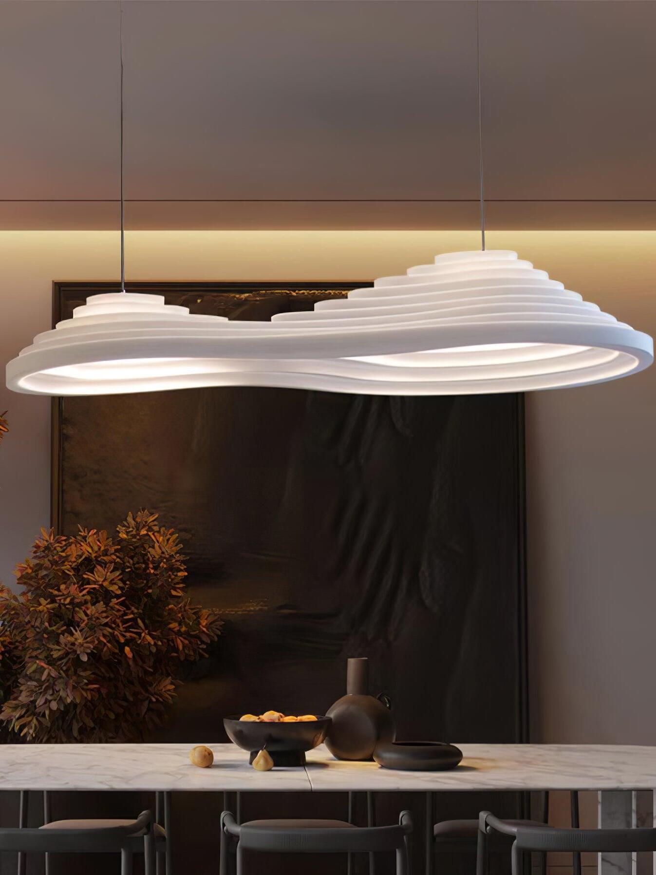 Elongated stepped pendant above a dining table with dark curtains and branches nearby.