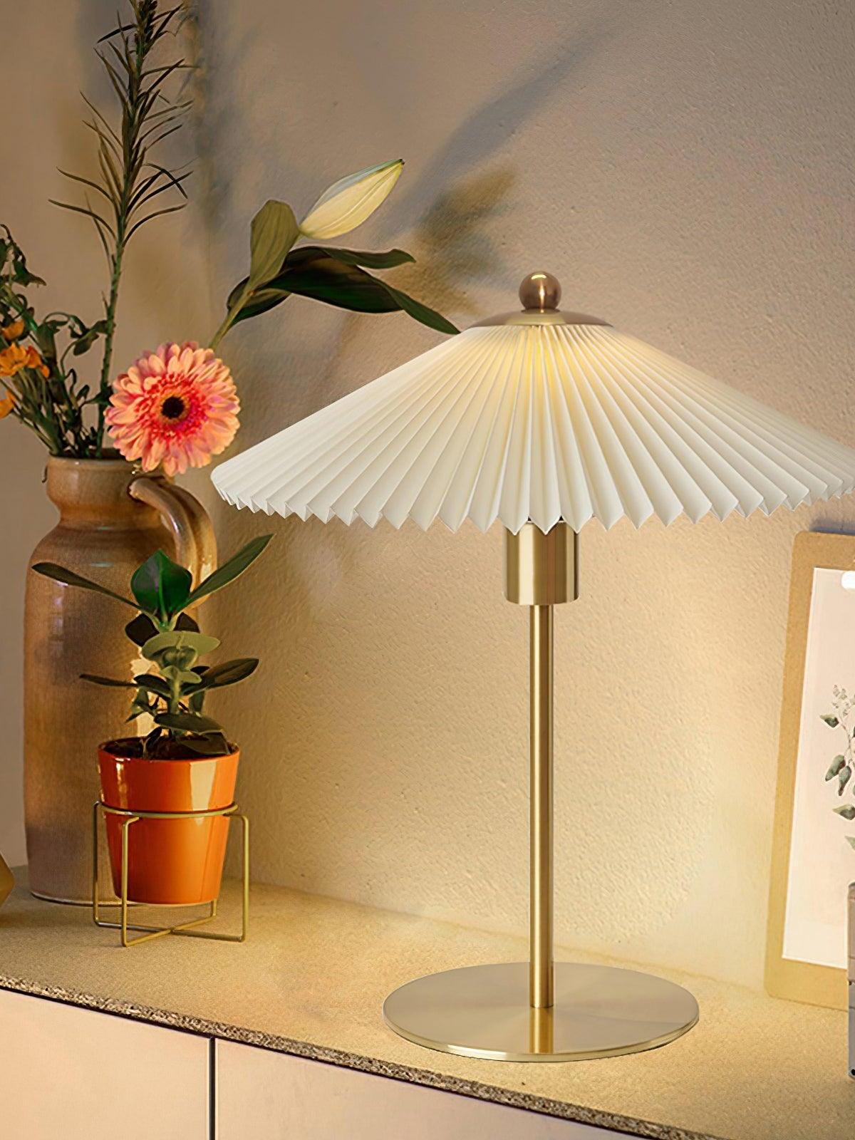 Lit pale pleated Perla lamp on a cabinet beside flowers and a small potted plant