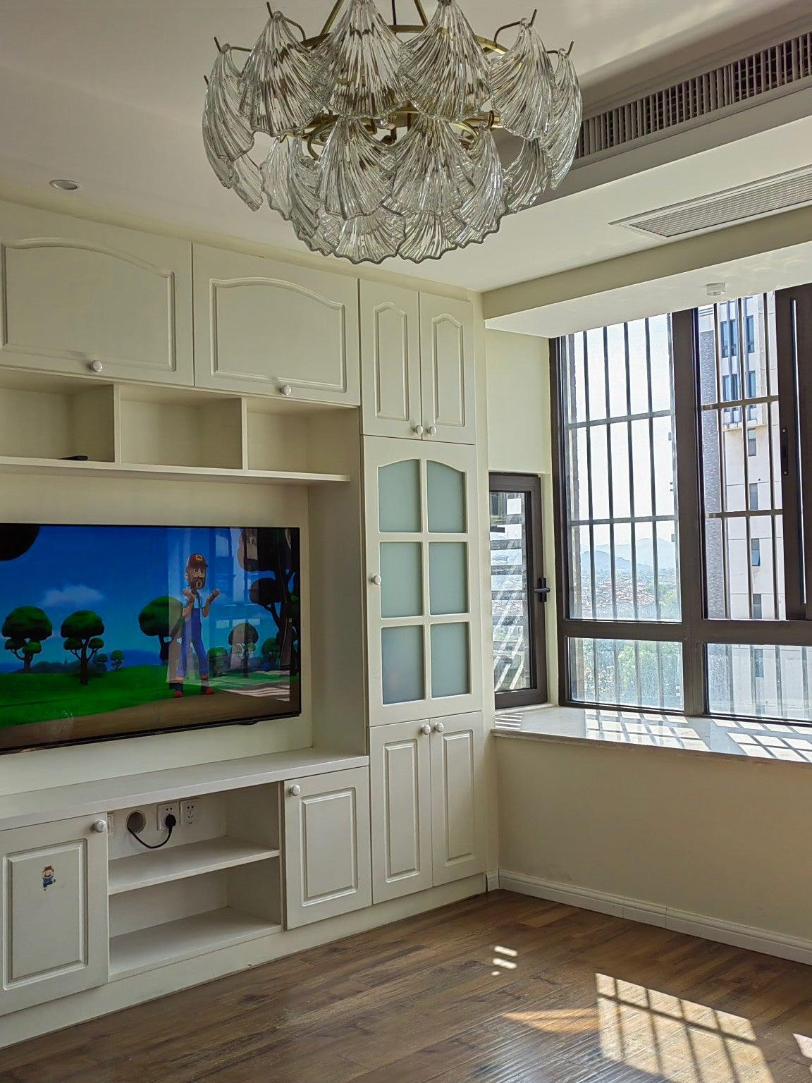 Unlit shell-panel fixture near a television, white fitted cabinets and a barred window