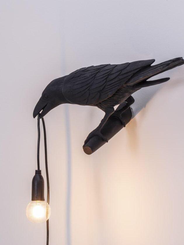 High-oblique black bird wall light showing feather texture, perch and hanging lit bulb