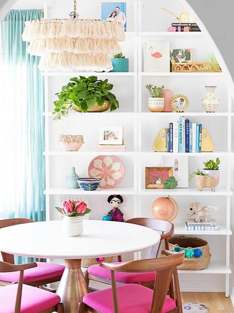 Light cream tassel chandelier over a white round table and pink-seated wood chairs, with plants and books on display shelves