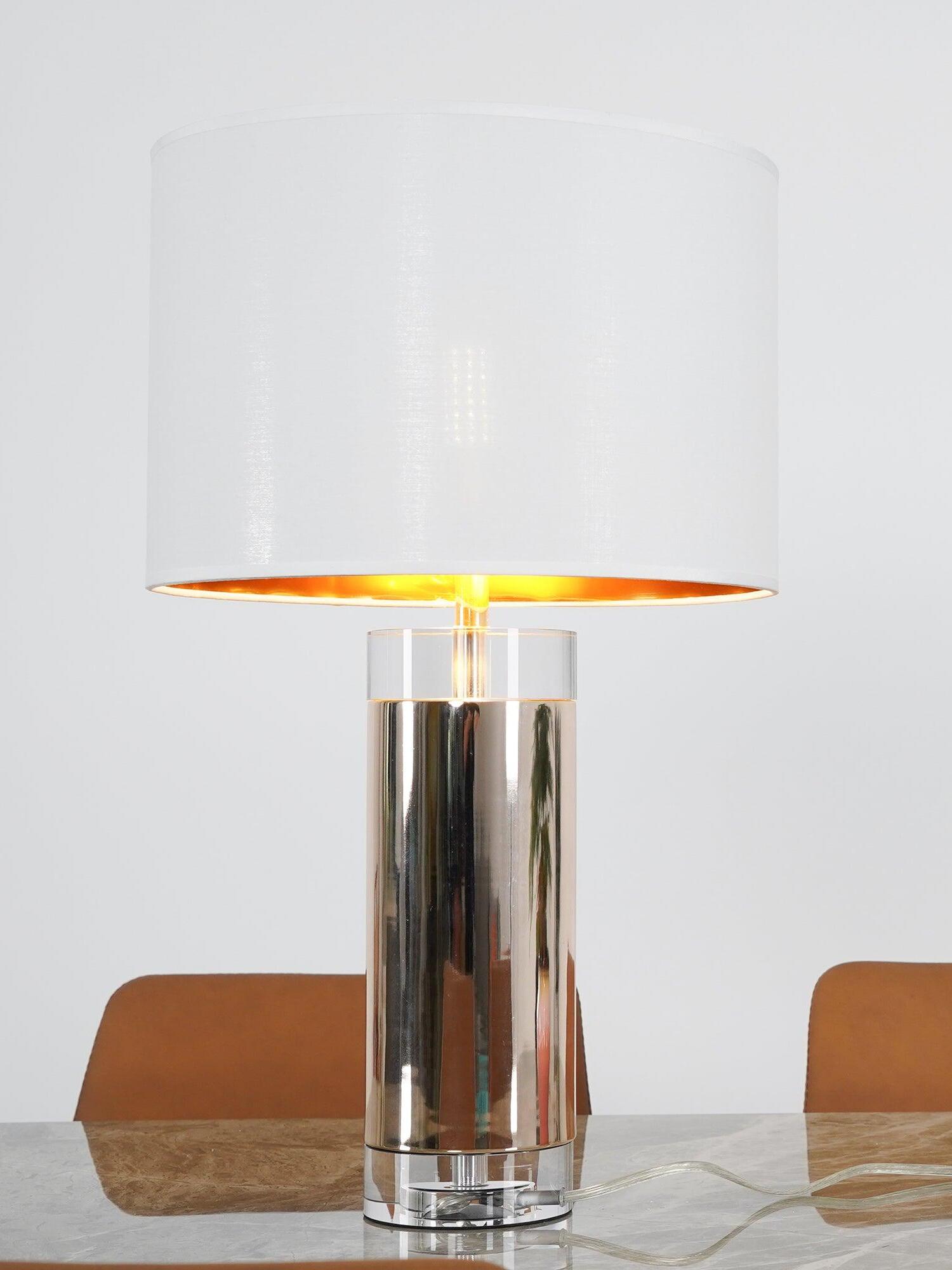 Lit white drum shade with warm gold-tone underside above reflective cylinder