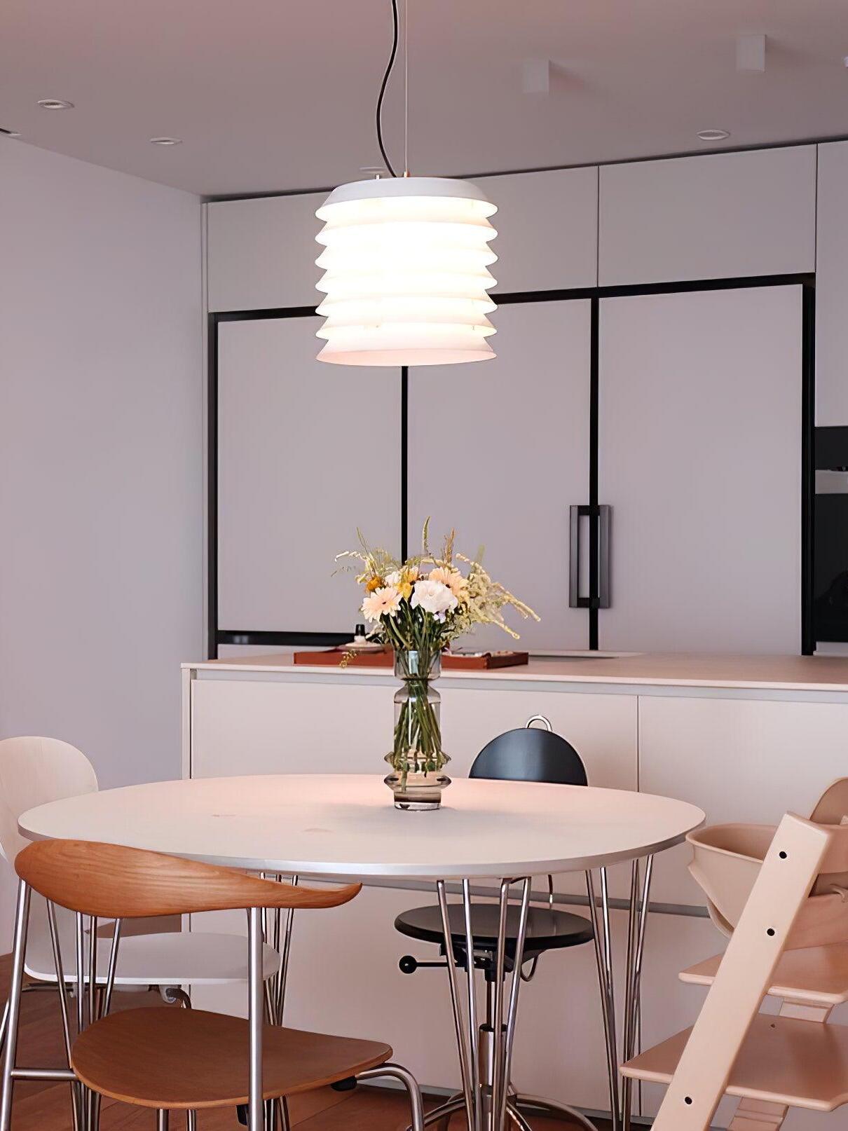 Lit white layered pendant above round table and vase in pale kitchen