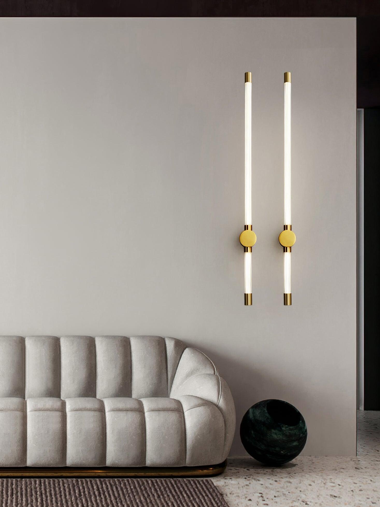 Pair of brass-tone vertical tube wall lights above a rounded sofa beside a dark green spherical ornament