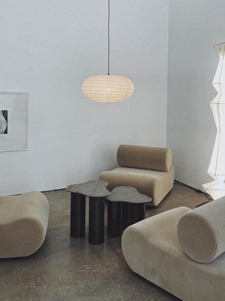 Pale flattened paper lantern on a dark cord above beige chairs and sculptural tables, with an angular paper lamp at right