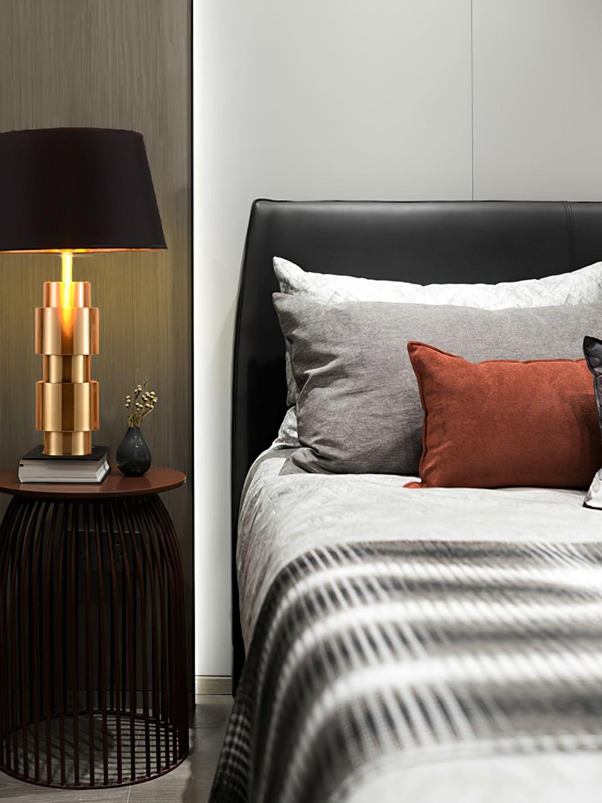 Glowing black-shade lamp on a round wire-frame bedside table beside gray bedding and a rust cushion