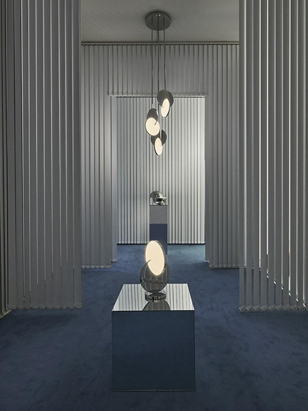 Chrome pendants on a shared round canopy beside a separate table lamp and mirrored plinth in a showroom