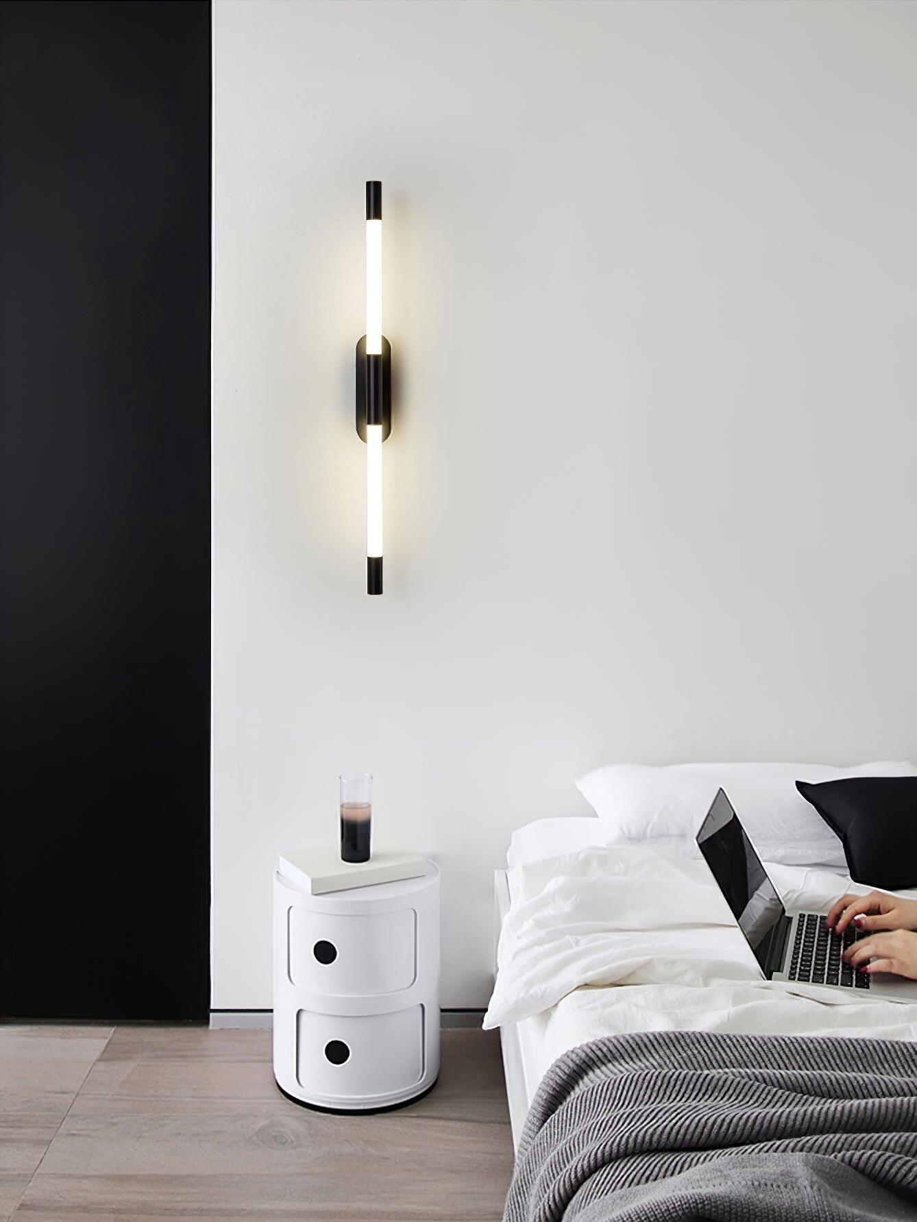 Vertical black Agnes wall light above a white cylindrical object beside a bed