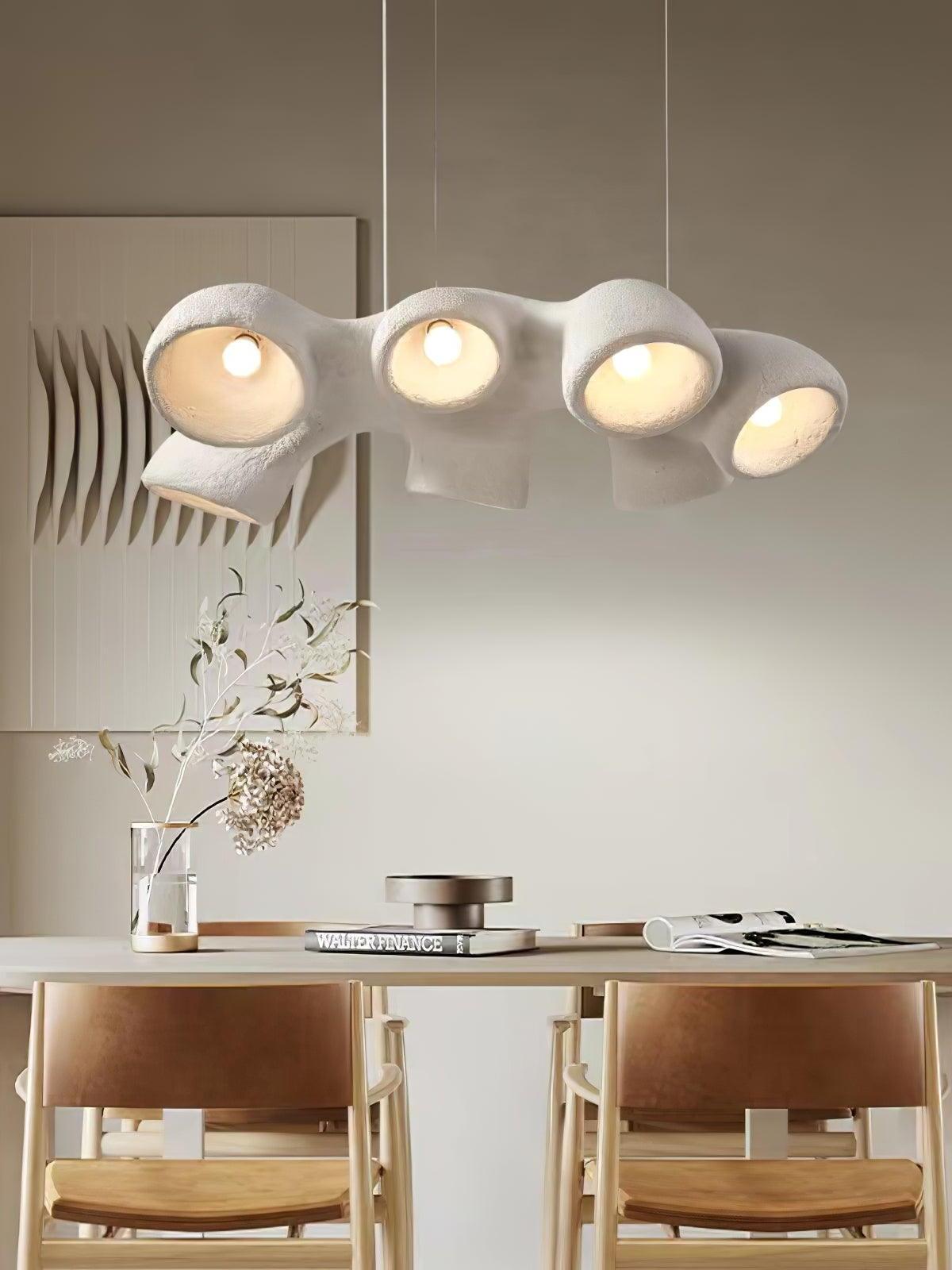 White branching pendant above a dining table with tan-backed chairs, vase branches, books and wall art